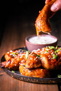Vegan Sticky Wings – plant-based chicken wings – One bitten one is held in one hand, others with dip on a plate.